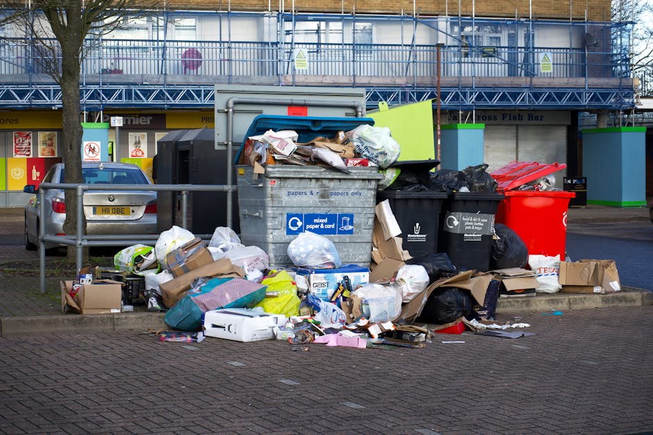 A collection of overflowing rubbish bins and scattered waste including cardboard boxes, plastic bags, paper, and packaging materials, situated on a paved area in front of a commercial building. The bins are of different sizes and colours—black, red, and grey—with some lids open, revealing mixed waste such as newspapers, plastic bottles, and disposable packaging. Nearby, loose waste materials lie on the ground, including flattened cardboard, plastic containers, and crumpled paper, contributing to a cluttered scene. Behind the waste piles, a metal railing separates the rubbish from a parking area where a dark grey car is parked. In the background, the building's façade features large glass windows and a blue scaffolding structure, indicating ongoing maintenance or renovation work. The environment appears to be an urban outdoor space, possibly a shopping centre or public plaza, with no active waste collection in process visible in the image. This scene exemplifies unmanaged waste accumulation that Rubbish Removal Wandsworth could assist with through private or alternative waste handling solutions.