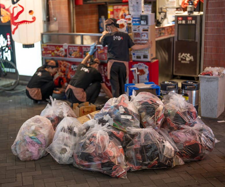 A scene outside a takeout restaurant shows multiple large clear plastic bags filled with assorted waste materials, including paper and plastic items, placed on a paved sidewalk. In the background, three workers dressed in black uniforms with brown aprons and gloves are actively engaged in clearing or organizing the waste, with two of them folding or stacking items near a small stand or delivery area. The scene is illuminated by ambient lighting, with surrounding elements such as vending machines, trash bins, and a red brick wall contributing to the urban environment. Visible on the right are two black canisters or containers, likely for waste collection, alongside a small metallic bin. The presence of large bags of mixed refuse and the workers' activities suggest a private or independent rubbish removal operation, compatible with services offered by companies like Rubbish Removal Wandsworth, in the context of event cleanup or site clearance at a busy commercial location.