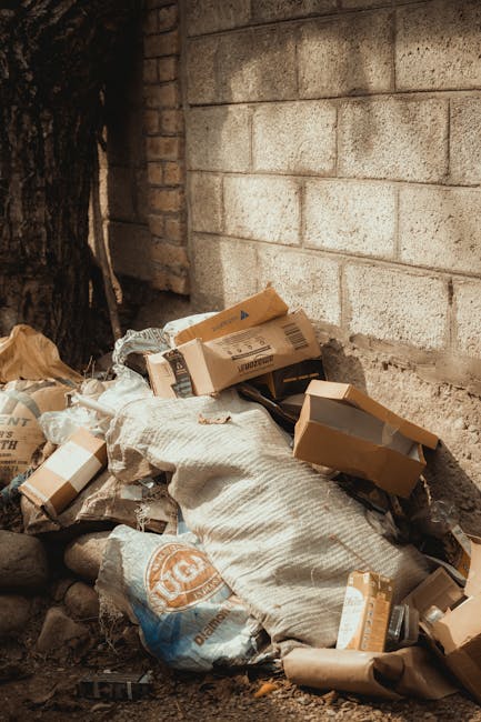 A collection of overflowing rubbish bins and scattered waste including cardboard boxes, plastic bags, paper, and packaging materials, situated on a paved area in front of a commercial building. The bins are of different sizes and colours—black, red, and grey—with some lids open, revealing mixed waste such as newspapers, plastic bottles, and disposable packaging. Nearby, loose waste materials lie on the ground, including flattened cardboard, plastic containers, and crumpled paper, contributing to a cluttered scene. Behind the waste piles, a metal railing separates the rubbish from a parking area where a dark grey car is parked. In the background, the building's façade features large glass windows and a blue scaffolding structure, indicating ongoing maintenance or renovation work. The environment appears to be an urban outdoor space, possibly a shopping centre or public plaza, with no active waste collection in process visible in the image. This scene exemplifies unmanaged waste accumulation that Rubbish Removal Wandsworth could assist with through private or alternative waste handling solutions.