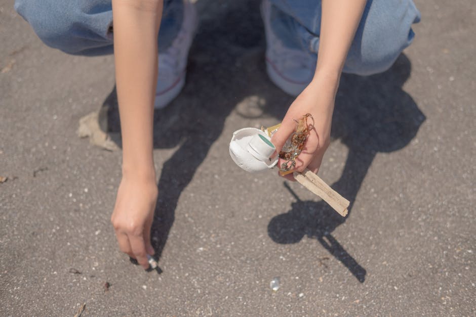 A person's hand is shown reaching towards the ground, gripping a small wooden stick with a white disposable cup attached to its top. The cup appears to be partly crushed or damaged, and there are some scattered small debris and dirt on the pavement. The person is wearing light blue clothing, with part of their arm and leg visible, and white shoes with a hint of pink detail. The background consists of a flat asphalt surface, suggesting an outdoor setting such as a driveway or street. This image illustrates the act of littering or the collection of rubbish, aligning with private waste removal or on-site clearance services provided by Rubbish Removal Wandsworth, especially in contexts that involve removing small-scale waste or debris from outdoor environments.