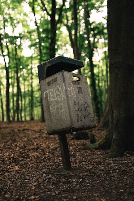 A worn, grey metal public mailbox with graffiti tags and scribbles on its surface, mounted on a slightly tilted black metal pole in a wooded area, surrounded by tall trees with green foliage. The mailbox appears to be in an outdoor setting, possibly near a park or nature reserve, with a forest floor covered in brown leaves and soil. The top of the mailbox has a hinged lid that is open, revealing the slot for depositing mail. The background features densely packed trees with trunks in various shades of brown and green, creating a natural backdrop that contrast with the utilitarian, slightly weathered appearance of the mailbox. The scene is illuminated by natural light filtering through the tree canopy, highlighting the texture of the graffiti and the rough finish of the metal. This image relates to the context of alternative waste handling or community-based rubbish disposal points, as it depicts a public collection point in a natural environment, relevant to rubbish removal services such as those provided by Rubbish Removal Wandsworth in the SW18 area near Wandsworth Common.