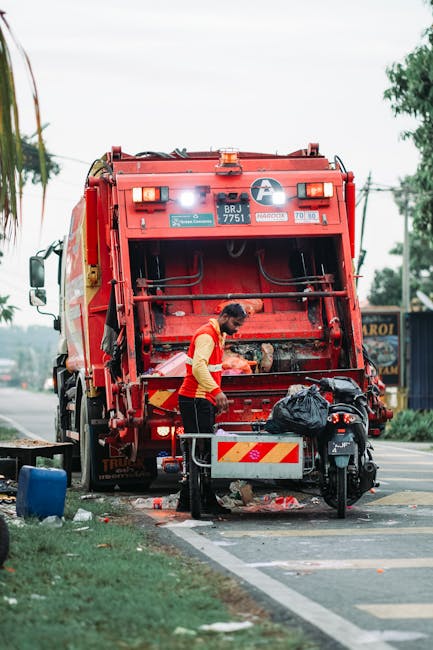 A waste collection worker wearing a high-visibility orange vest and dark trousers is operating a large red rubbish collection vehicle on a street. The vehicle's rear hopper is open, revealing mechanical components designed for compacting waste. The worker is leaning slightly forward, handling black plastic garbage bags that are placed on a small platform attached to the vehicle, possibly preparing them for loading or sorting. The scene is outdoors during daylight hours, with a partly cloudy sky, and the vehicle is positioned at the edge of a paved road with some debris, small waste items, and discarded refuse visible on the ground nearby. To the left, a small patch of grass with a blue container or bin is present, and in the background, there are trees, power lines, and a few roadside structures, including signage for local businesses. The setting suggests a typical urban or suburban environment where private waste collection services, such as those provided by Rubbish Removal Wandsworth, are in operation for alternative rubbish removal or on-site clearance, supporting off-form municipal waste collection. The scene emphasizes the active process of rubbish handling and disposal in a professional waste management context. The image visually underscores the practical aspects of rubbish removal services dealing with domestic or commercial waste outside of standard municipal collection systems.