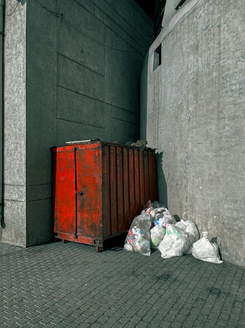 A red metal waste container with a weathered finish and visible rust spots is positioned against a dark green building facade on the left and a light gray concrete wall on the right. The container has a horizontal slatted design on its sides and is partially opened at the top, though its contents are not visible. Several white plastic bags filled with rubbish are clustered on the ground beside the container, some resting on the textured brick pavement and others leaning against the container. The bags appear to contain mixed waste, including visible plastic bottles and packaging. In the background, a small rectangular window or ventilation opening is embedded in the concrete wall, and the scene is lit with natural daylight, casting subtle shadows on the pavement. This setting suggests a private waste disposal or alternative rubbish collection point, aligned with the services offered by Rubbish Removal Wandsworth for local residents needing on-site clearance or non-standard rubbish removal solutions.